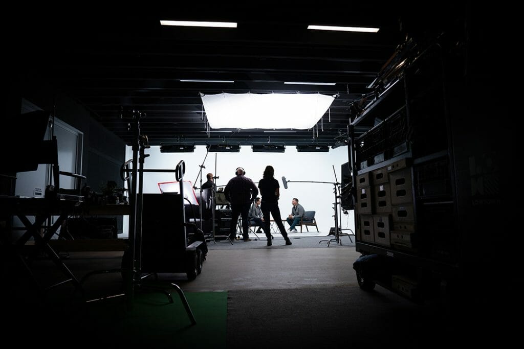 Behind the scenes photo of a set with camera equipment and lighting surrounding two men talking in chairs.